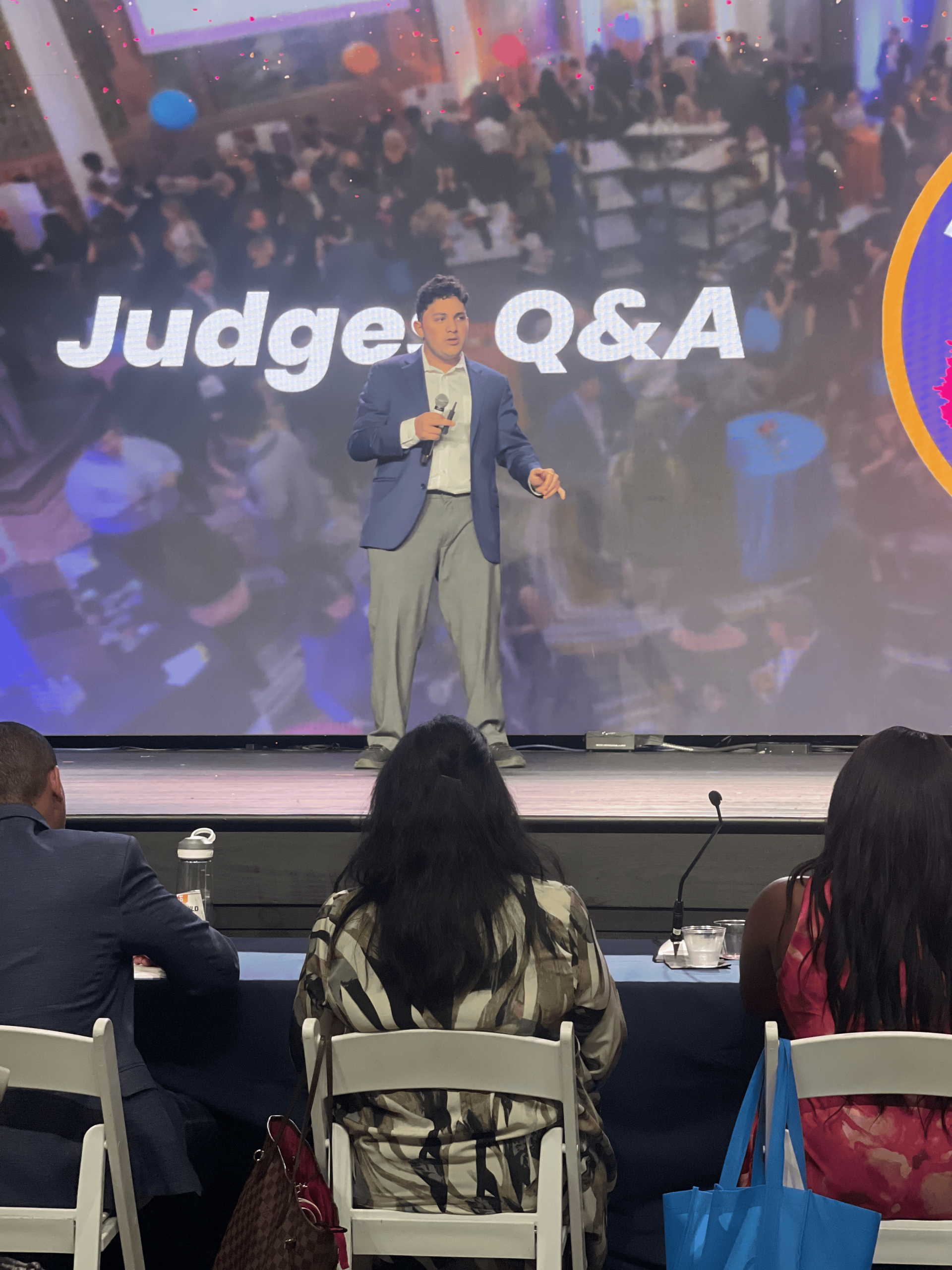 IMG_7444 A man who is named Evan A. Khanna in a blue suit stands on stage holding a microphone, speaking to an audience. The screen behind him reads “Judge Q&A.” Three people sit facing him in the foreground.